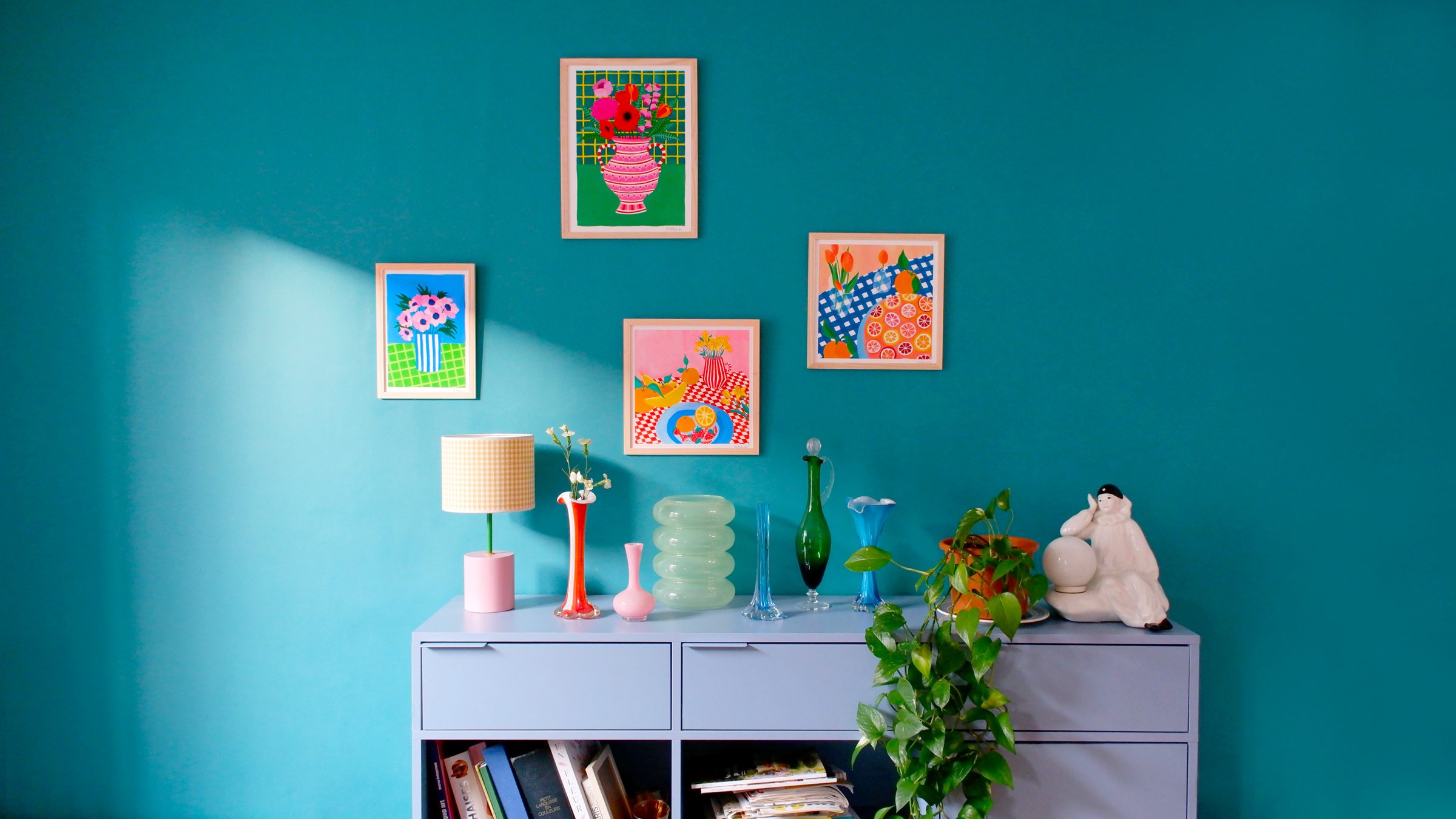 Quatre tirages Kittana encadrés sur un mur bleu canard, au-dessus d'un meuble bleu lavande avec des vases colorés
